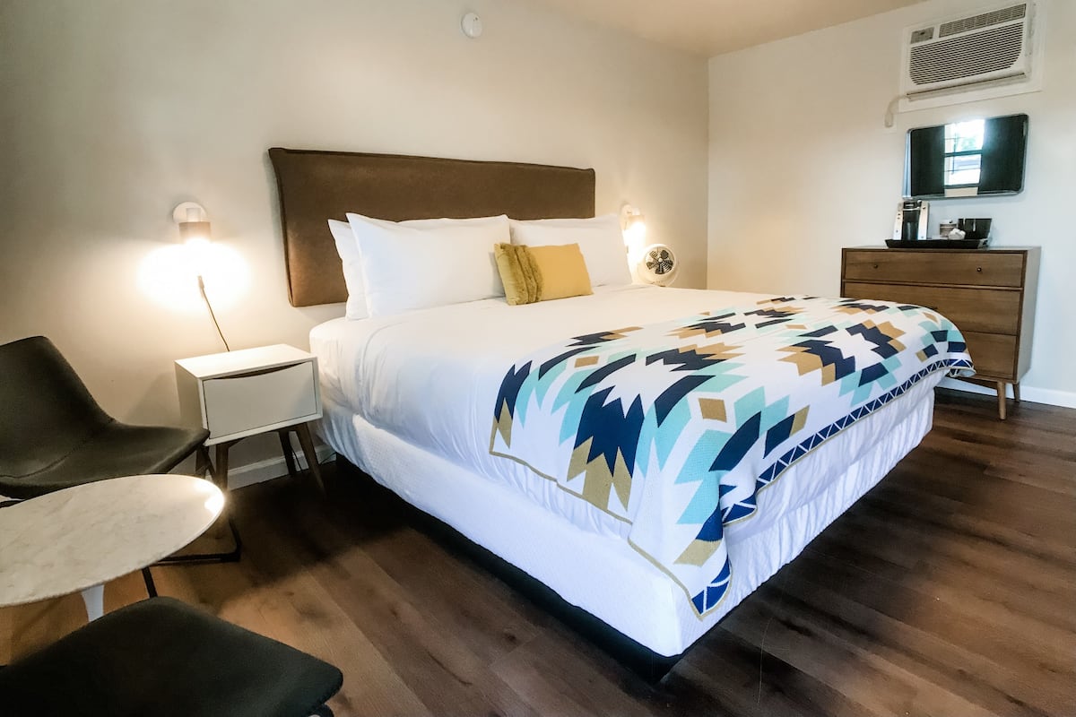 A hotel room with a bed, bedside table, chair, dresser, and a small table.