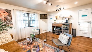 A modern living room with a wooden floor, a glass coffee table, a sofa, a patterned rug, a TV, and a kitchen area with a coffee machine and shelves. - Stonewall Motor Lodge (Stonewall)