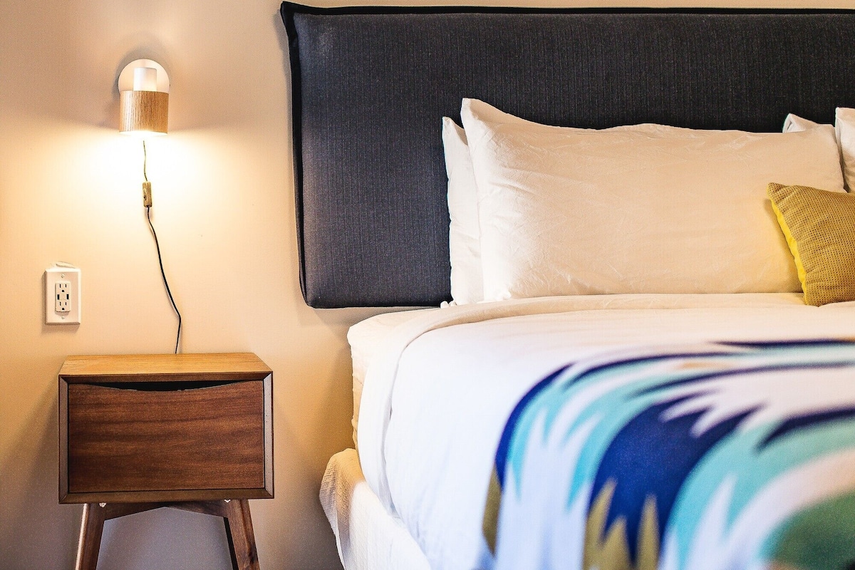 A neatly made bed with white and beige bedding, a dark headboard, and a yellow pillow. A bedside lamp is on the wooden nightstand beside the bed.