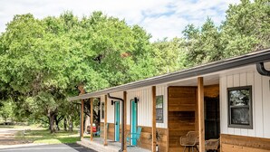 A motel with a covered outdoor seating area, a parking lot, and a large tree in the background. - Stonewall Motor Lodge (Stonewall)