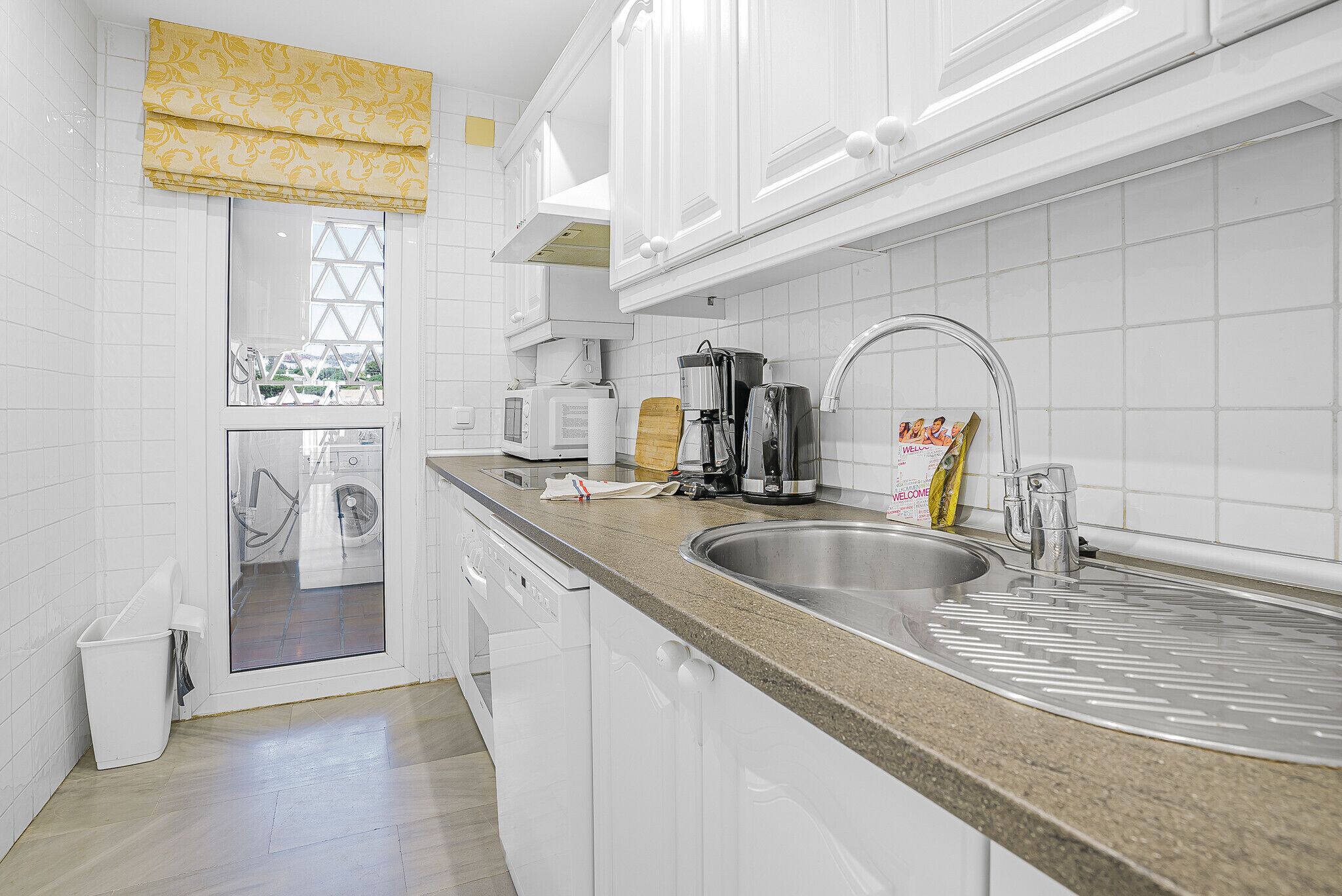 A modern kitchen with white cabinets, a granite countertop, a stainless steel sink, and a window with a yellow valance.