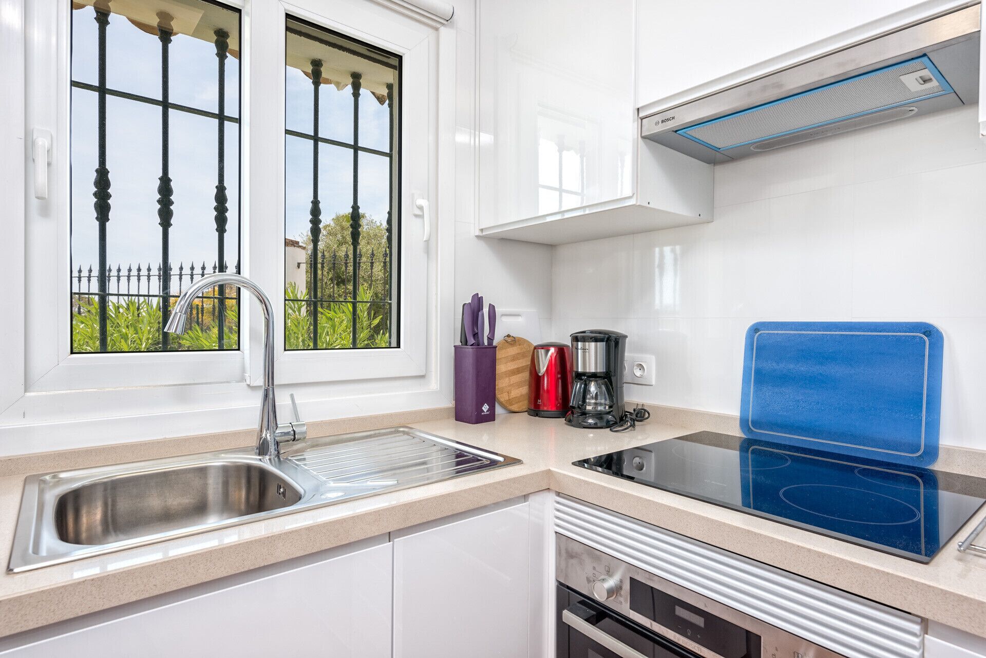 A modern kitchen with a stainless steel sink, a Bosch coffee maker, a blue induction cooktop, and a window with a view of greenery.