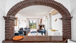 A modern living room with a stone archway, a sofa, a dining table set for two, and a view of the outdoors. - F04 Bahia Playa - Apartment (Mijas Costa)