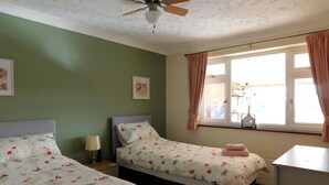 A bedroom with two beds, a ceiling fan, a window with curtains, and a bedside table with a lamp. - Sky View -  Enjoy panoramic roof top views from the sunny conservatory or outside deck. (Sheringham)