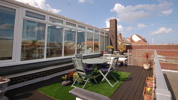 A modern outdoor patio with a glass enclosure, a wooden deck, and a seating area with chairs and tables. - Sky View -  Enjoy panoramic roof top views from the sunny conservatory or outside deck. (Sheringham)