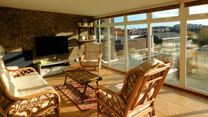 A spacious living room with a brick wall, a flat-screen TV, wicker furniture, and a large sliding glass door offering a view of the neighborhood. - Sky View -  Enjoy panoramic roof top views from the sunny conservatory or outside deck. (Sheringham)