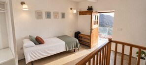 Un dormitorio con cama, mesita de noche, banco de madera, estante en la pared y puerta que da a un balcón con vistas a la montaña.