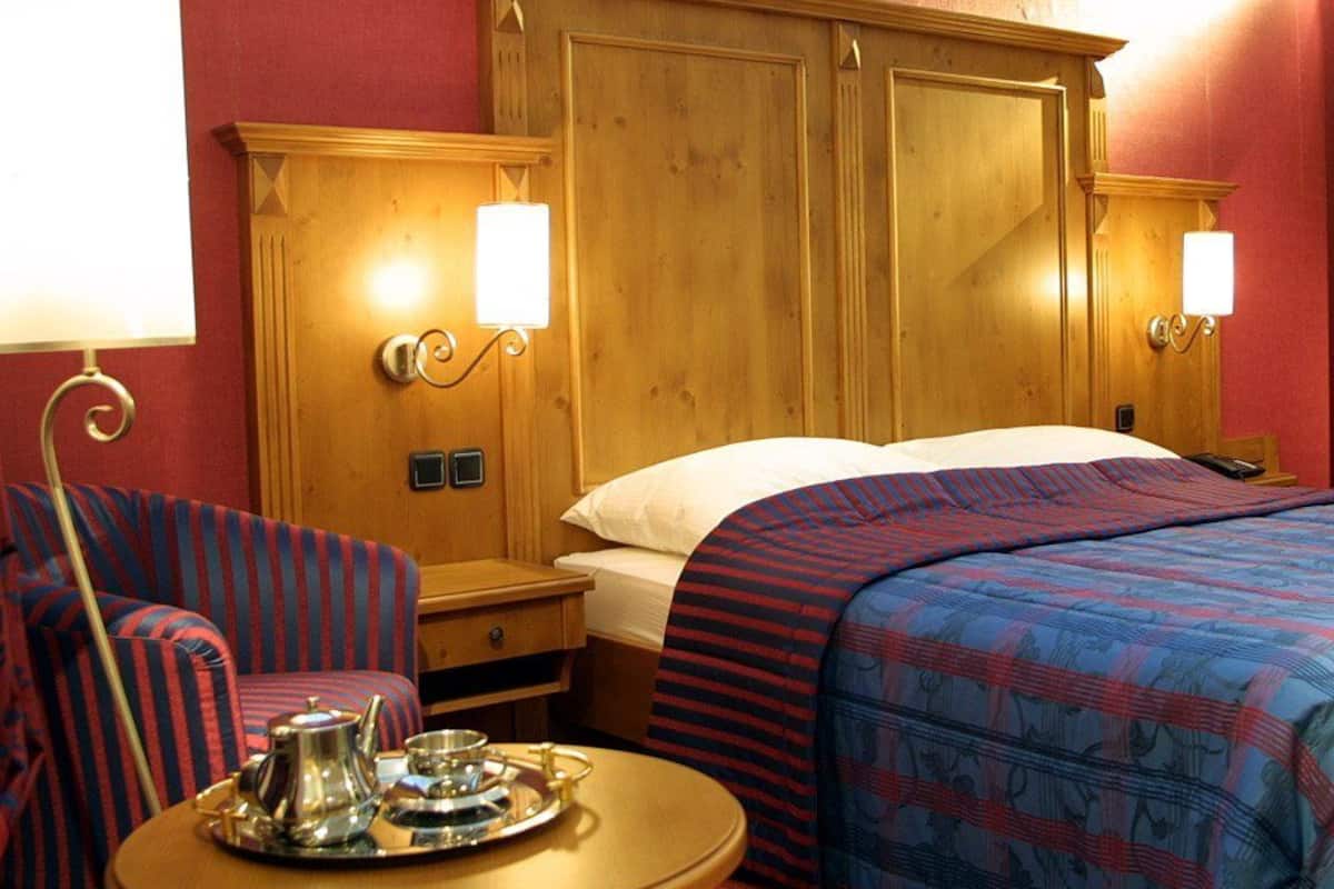 A hotel room with a wooden headboard, two bedside lamps, a striped armchair, a round table with a tray, and a patterned bedspread.