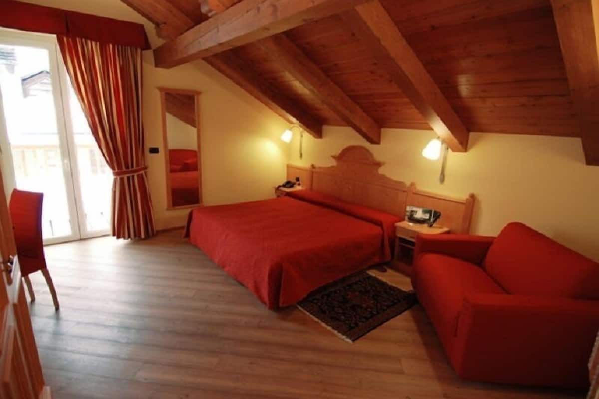 A bedroom with a wooden ceiling, a bed with a red bedspread, a nightstand, a red sofa, and a window with curtains.