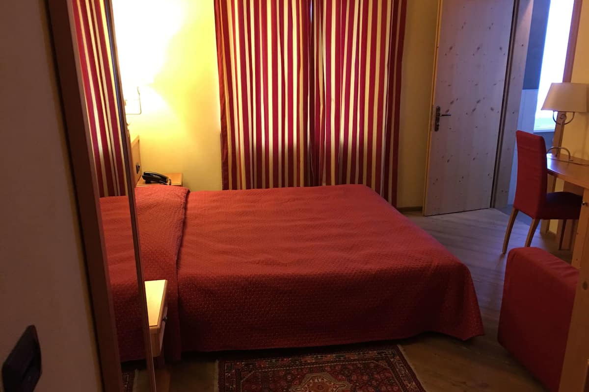 A small, cozy room with a bed, a chair, a desk, and a patterned rug.