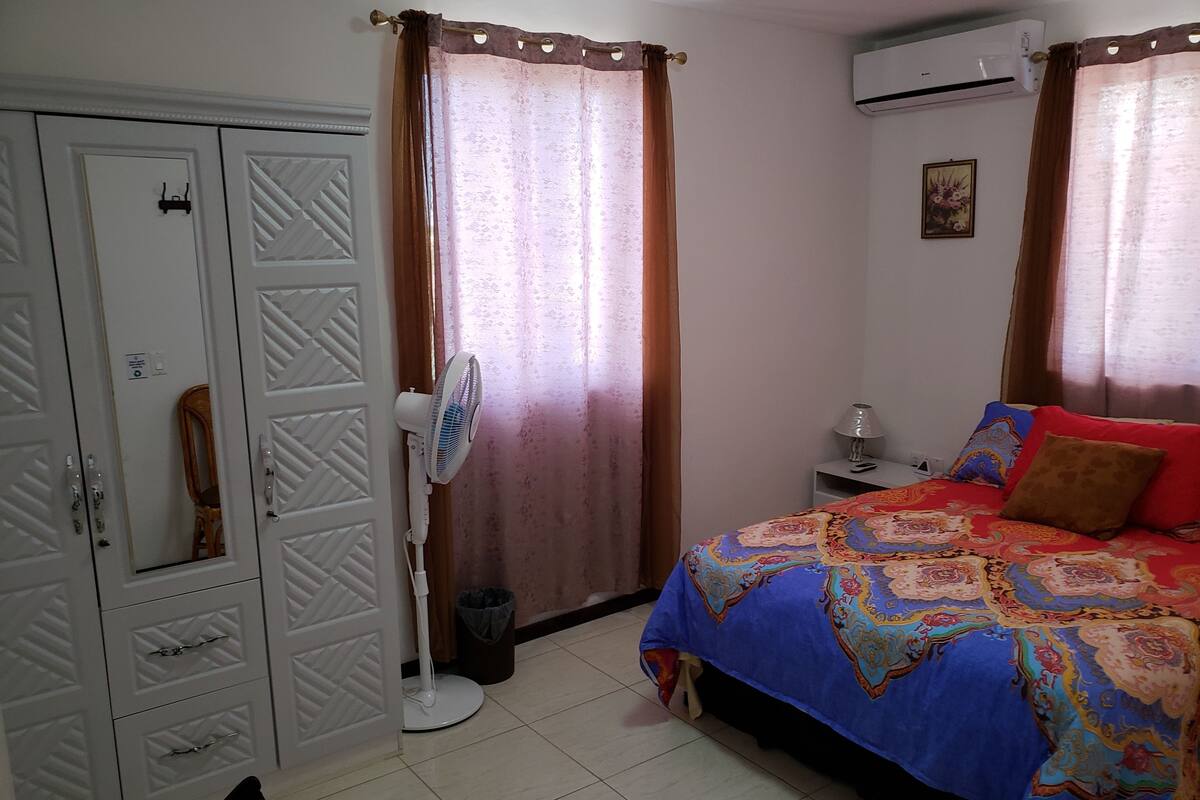 A bedroom with a bed, a wardrobe, a standing fan, and an air conditioning unit.