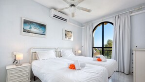 Et hotelværelse med to senge, ventilator, aircondition, et natbord med lampe og et stort vindue med gardiner.
