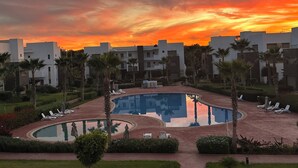 A resort with a swimming pool, palm trees, and modern buildings during sunset. - High standing apartment PERLA SAIDIA 3min walk from the beach (Saïdia)