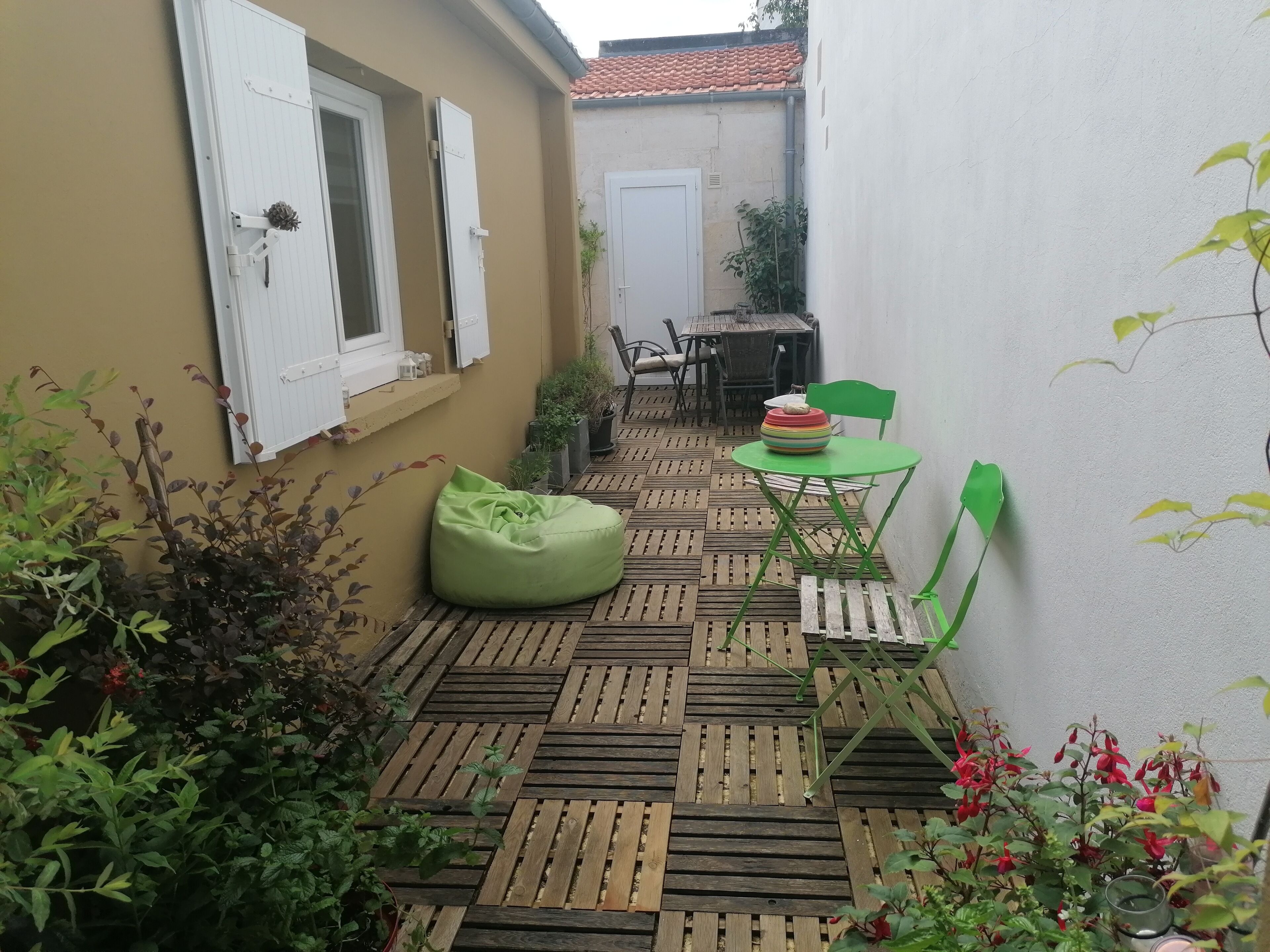 Un pequeño patio con suelo de madera, sillas verdes, una mesa y plantas en macetas.