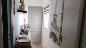 Ein schmaler Flur mit einem Kühlschrank, einer kleinen Pflanze und einem dekorativen Gegenstand auf einem Regal. Im Hintergrund ist eine Tür mit Fenster zu sehen.