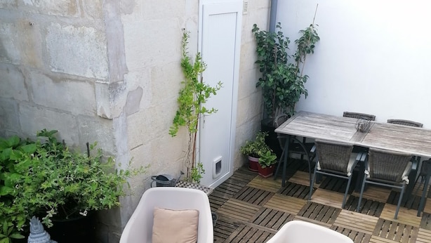 Eine Terrasse im Freien mit Holztisch, zwei weißen Plastikstühlen, einem kleinen Tisch und Blumentöpfen.