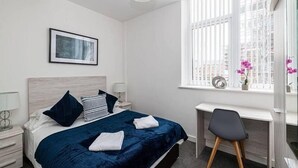 A hotel room with a bed, a desk, a window with blinds, a picture on the wall, and a nightstand with a lamp.