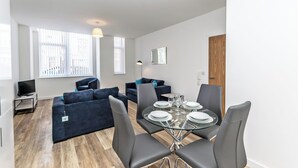 Un salon moderne avec une table à manger en verre, des canapés sombres et un téléviseur posé sur un meuble.