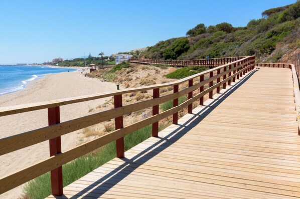 A wooden boardwalk along a beach with a clear view of the ocean and a hilly landscape in the background. - B07 Caserio - Apartment close to the beach (Sitio de Calahonda)
