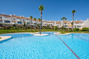 A swimming pool with clear blue water, surrounded by white buildings with red-tiled roofs and palm trees.