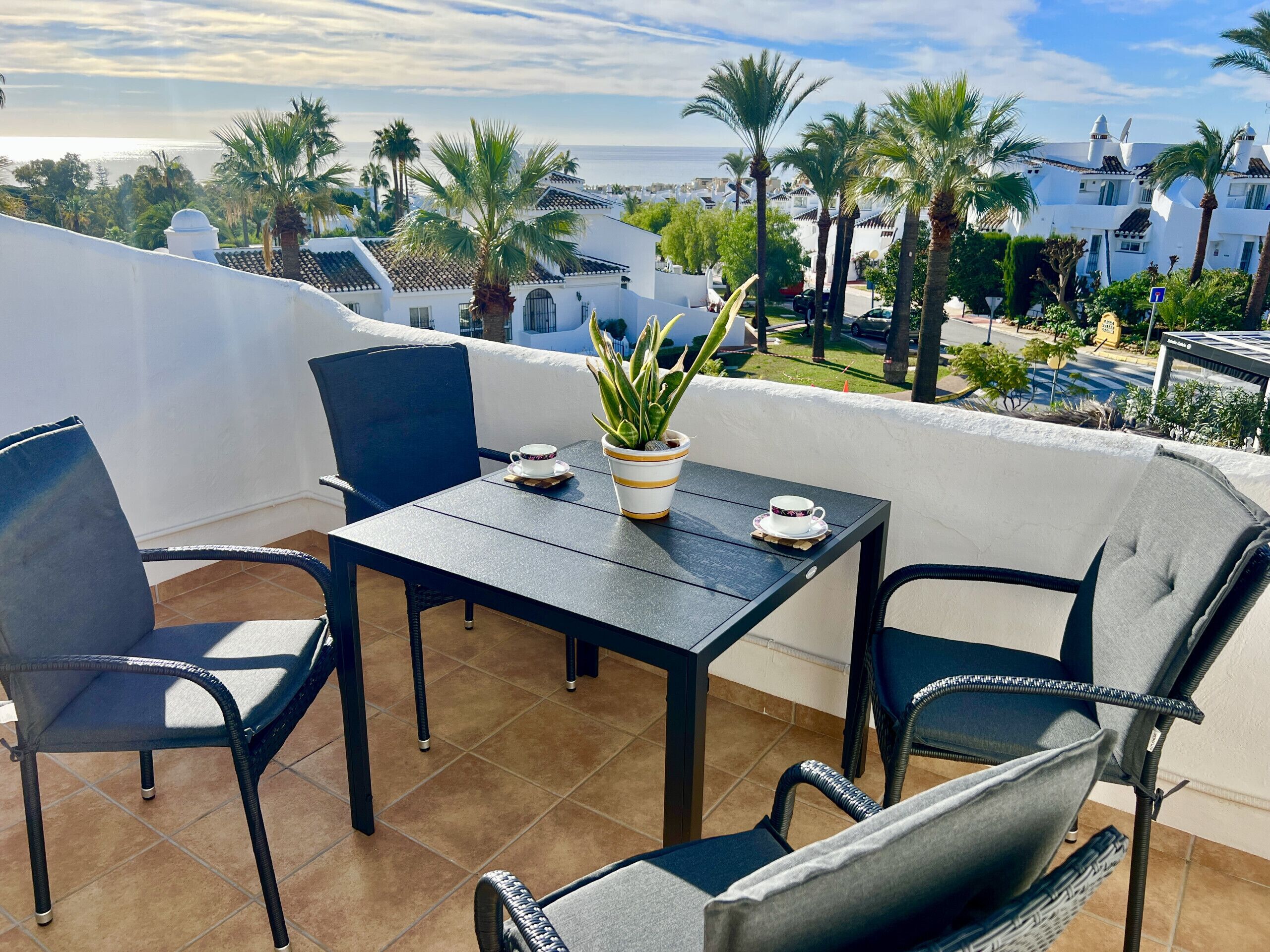 Un balcon avec une table et des chaises, un pot de fleurs et une vue sur des palmiers et des immeubles.