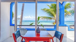 Un coin repas avec une table et des chaises, un vase avec une plante, et une vue sur l’océan et les palmiers, visible à travers de grandes fenêtres.
