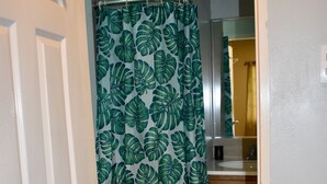 Un baño con cortina de ducha estampada con hojas, una alfombra verde y armarios de madera.
