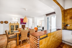 Una cocina moderna con salpicadero de ladrillo, un comedor con mobiliario de mimbre y vistas al salón a través de un vano abierto.