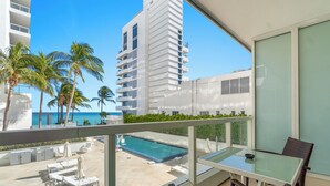 A balcony with a glass table and wicker chairs, overlooking a swimming pool and palm trees.