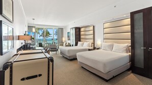A hotel room with two beds, a desk, a chair, a suitcase, and a large window with a view of palm trees and the ocean.