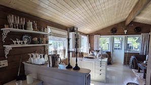 A cozy cabin interior with wooden walls, a kitchen area with white cabinets, and a dining table set with glassware. - High standard cabin with view, hiking and fishing Ocean boat rental possible (Raudsand)