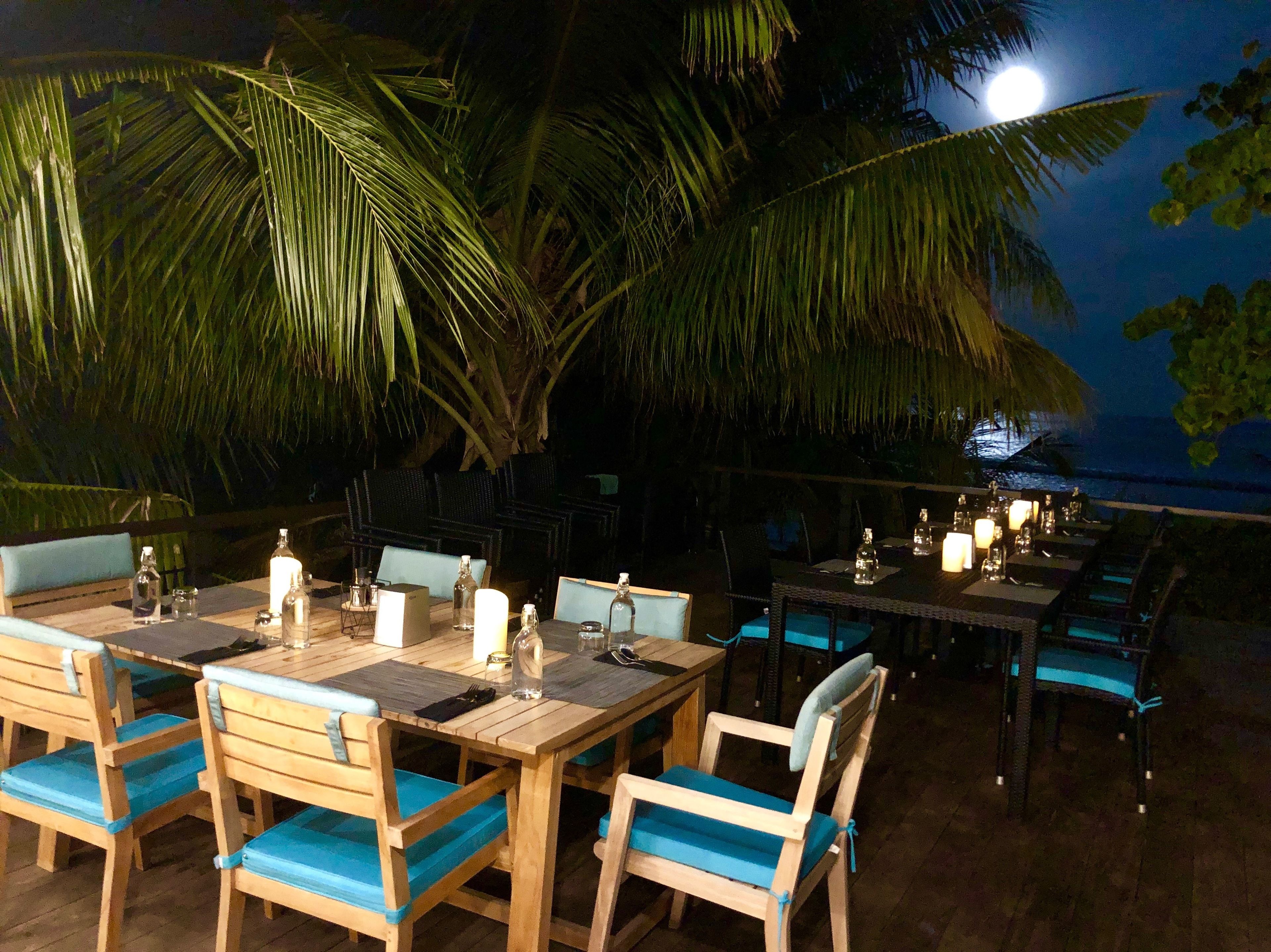 Un espacio exterior para comer con mesas y sillas de madera, velas y vista al océano bajo una luna llena.