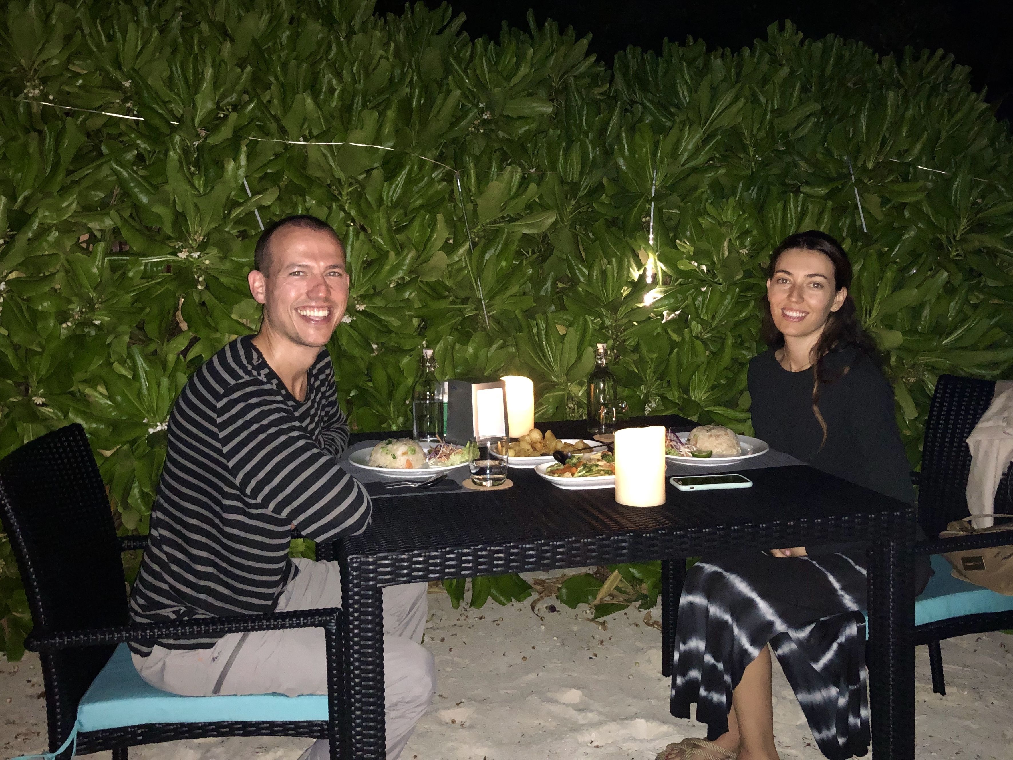 Two people seated at an outdoor table with plates of food, surrounded by greenery and string lights.