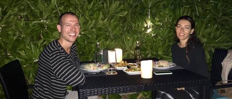 Two people seated at an outdoor table with plates of food, surrounded by greenery and string lights.