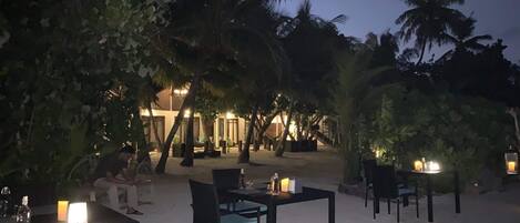 Un área de comedor al aire libre con mesas y sillas, rodeada de palmeras y un edificio al fondo.
