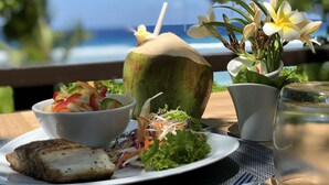 Una comida con pescado a la parrilla, ensalada y una bebida de coco sobre una mesa con vista al océano.