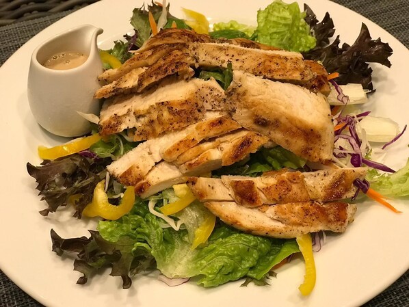 A plate of grilled chicken with mixed greens and yellow bell peppers, accompanied by a small bowl of dressing.