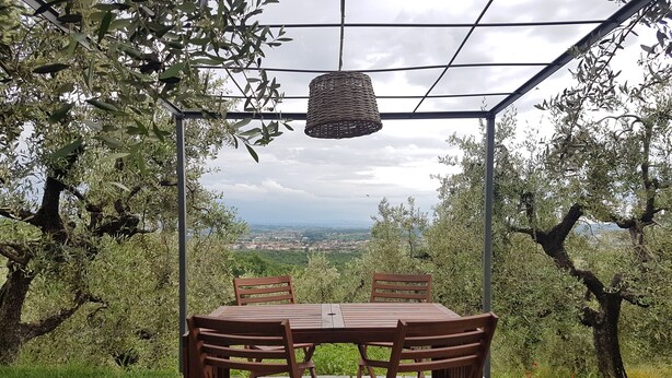 Ein Außenbereich zum Sitzen mit Tisch und Stühlen, umgeben von Olivenbäumen und Blick auf eine ferne Stadt.
