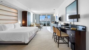 Moderni hotellihuone, jossa on suuri sänky, ruokailutila baarituoleineen, taulutelevisio ja näkymä kaupunkiin.