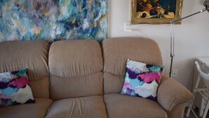 Een bruine bank met twee kleurrijke kussens, een schilderij van fruit en groenten, en een witte lamp.