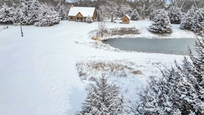 A snow-covered landscape with a pond, a stone house, and a wooden cabin.