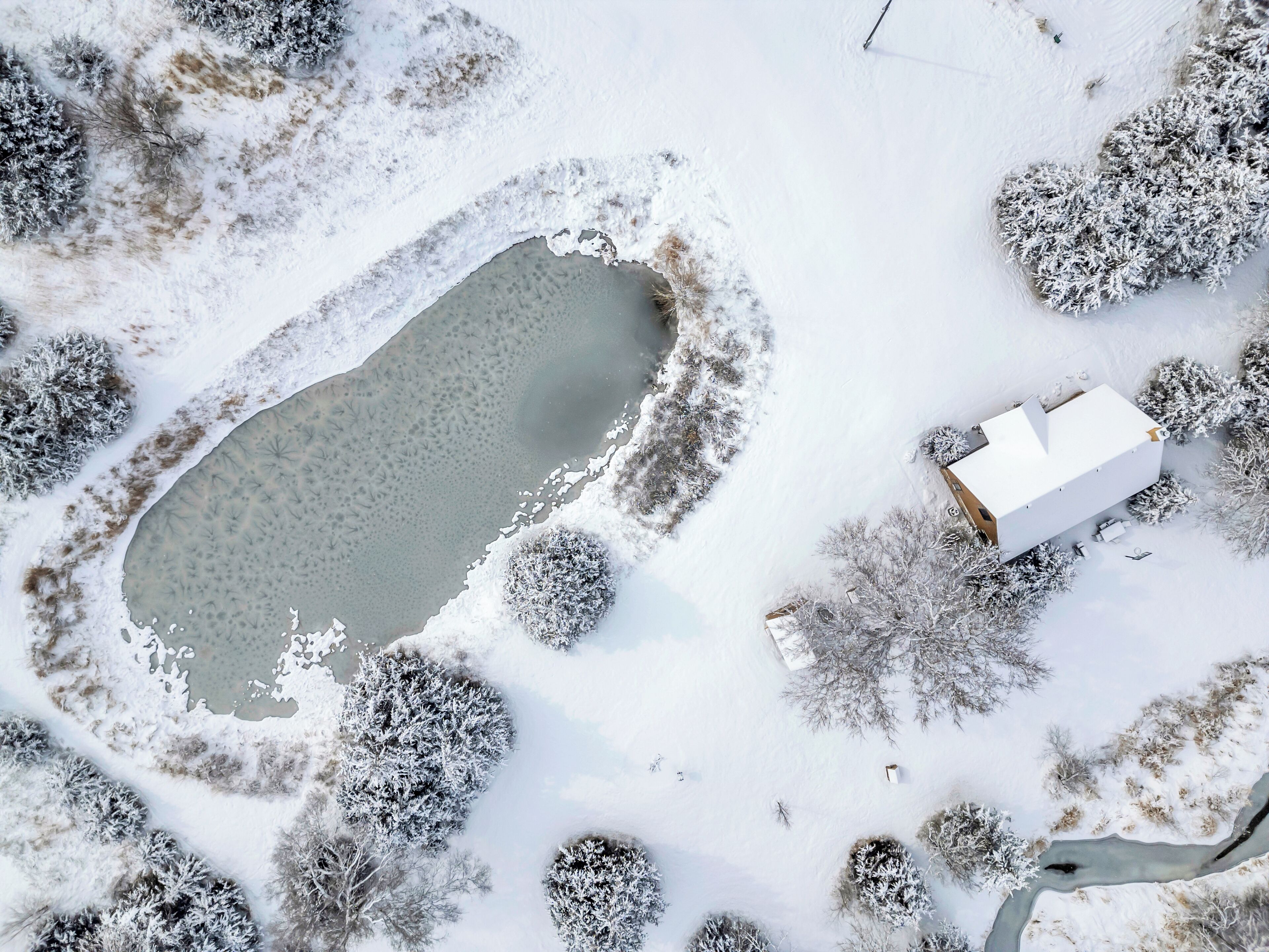 An aerial view of a snow-covered landscape with a pond, a white building, and surrounding trees.