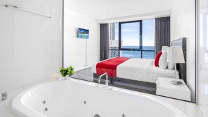 A modern hotel room with a large bathtub, a bed with red and white bedding, a view of the ocean, and a small plant on the side. - Circle on Cavill Ocean & River Views (Surfers Paradise)