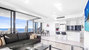 A modern living room with a sofa and a glass coffee table, a dining area with chairs, a kitchen with white cabinets, and a large window offering a city view. - Circle on Cavill Ocean & River Views (Surfers Paradise)