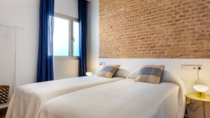 Een kamer met twee bedden, een bakstenen muur, een raam met blauwe gordijnen en een nachtkastje met een lamp.