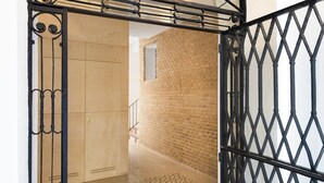 Een zwart ijzeren hek met een geometrisch ontwerp, leidend naar een hal met een vloerbedekking met patroon en een bakstenen muur.