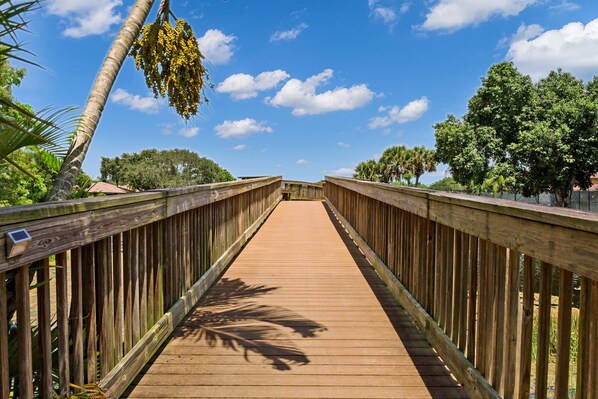 Un paseo marítimo de madera que lleva a un puente, con cielo despejado y árboles al fondo.