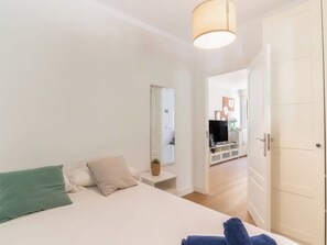 A bedroom with a bed, a bedside table with a lamp, a mirror, and a view into the living area.