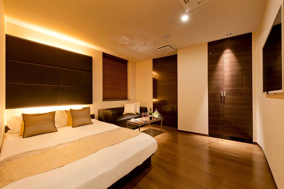 A modern hotel room with a large bed, a dark headboard, a wooden wardrobe, and a small seating area with a table and chairs.
