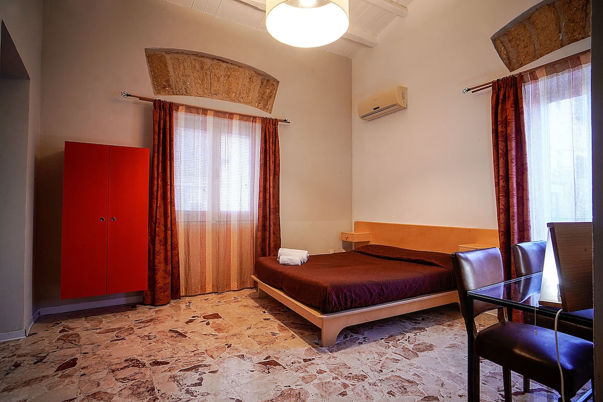 A bedroom with a bed, a red wardrobe, a desk with a chair, and a window with curtains.